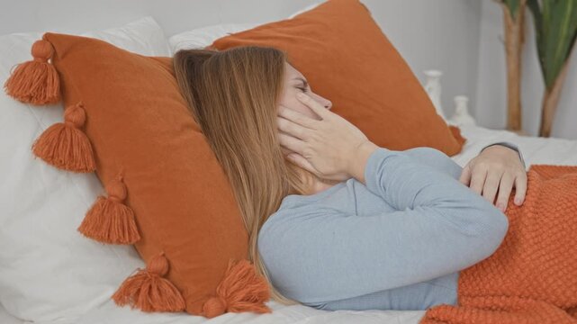 Young woman lying in bed with hand on cheek indicating toothache, covered by orange blanket, leaning on tassel pillow in building; toothache pain.