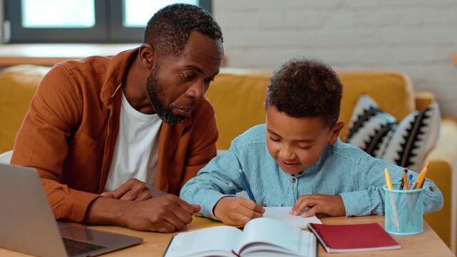 A man with dark skin and a boy work together at a table in a cozy living room. The boy writes in a notebook while the man guides him, creating a warm learning environment.