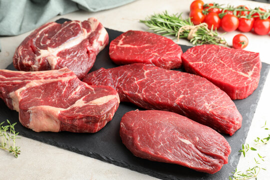 Different raw beef steaks, tomatoes and herbs on light table, closeup