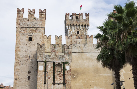 View of the historic Scaligero Castle featuring medieval stone towers with swallowtail battlements and lush palm trees in Italy.
