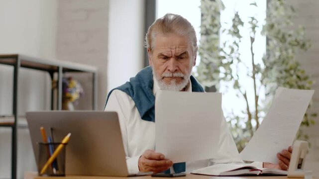 A senior man with gray hair and a beard sits at a desk, looking at papers while using a laptop. He is focused and engaged in his work in a well-lit office.