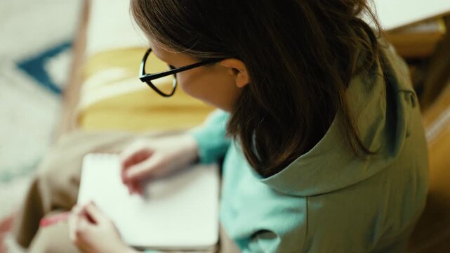 Smart Little Girl in Big Glasses Writing in Notebook while Sitting on Couch with Stack of Books &mdash; Cinematic 4K 10-bit
