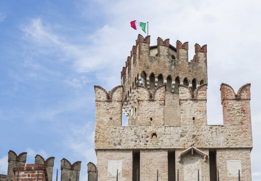 View of Scaligero Castle featuring the medieval stone tower with swallow-tail merlons and the Italian flag waving against a blue sky in Italy.