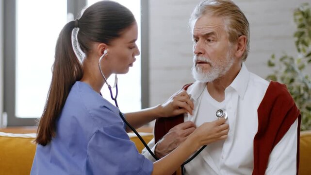 A woman in blue scrubs checks the heart rate of an older man in a white shirt. They are in a bright room with a couch. The woman uses a stethoscope, showing care and attention.