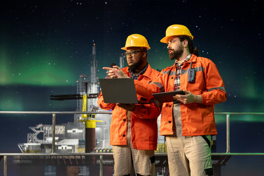 Offshore platform engineering team using laptop to control subsea systems remotely at night. Professionals monitoring anomaly detection using notebook on drilling derrick
