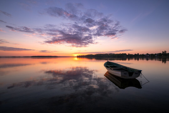 View of a small wooden boat floating on the calm surface of a lake during a vibrant sunset with purple clouds Piaski, Warmian-Masurian Voivodeship, Poland.