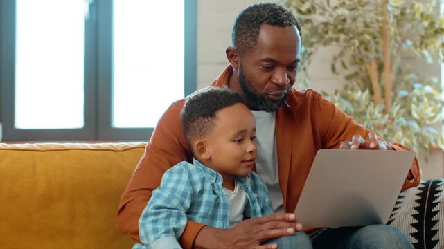 A man with dark skin sits on a couch with his young son. They are looking at a laptop together, enjoying a moment of learning and connection in their cozy living room.