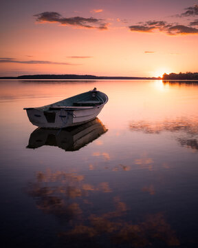 View of a small rowing boat floating on a calm lake during a vibrant sunset with orange clouds and reflections in Piaski, Warmian-Masurian Voiv, Poland.