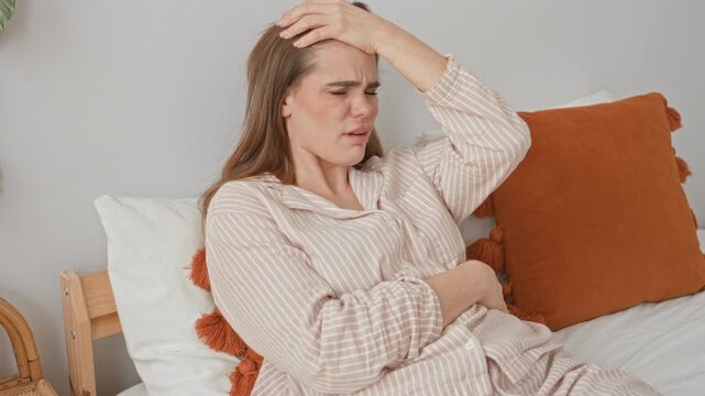 Woman clutching stomach and hand on forehead on bed inside building with orange cushion and striped pajamas, tense expression; discomfort nausea.