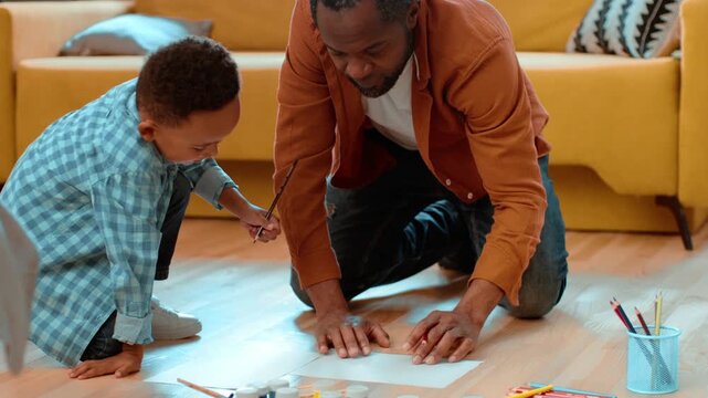 A man with dark skin and a young boy are painting together on the floor. They are surrounded by art supplies and are sharing a fun moment full of creativity in their living room.