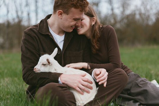 A couple in casual clothing cuddle a white goat kid in a sunny field, creating a warm rural moment. Lifestyle portrait showing love, affection, and the bond between humans and animals on a meadow.