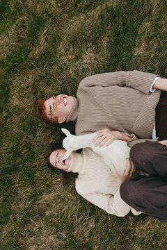 Cheerful couple outdoors, lying on grass with a playful goat kid, cozy knit sweaters, warm natural tones, relaxed smiles, candid moment of connection and countryside vibes