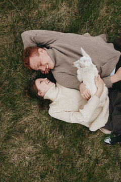 Two people lying on grass outdoors smiling and cuddling a white goat kid. A warm cozy moment capturing affection, nature, and simple farm life with soft light and a relaxed mood.