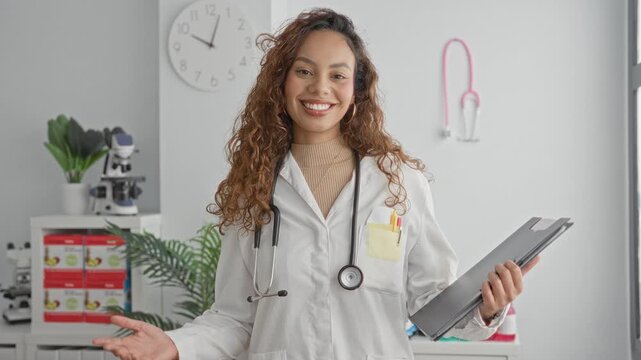 Woman doctor holding clipboard and stethoscope, smiling with open hand gesture in medical building; care confidence.