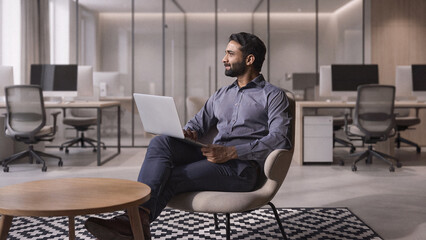 Young thoughtful bearded smart indian professional business man executive looking away sitting on chair in modern office lobby using laptop, thinking of new ideas, planning project, working online. © insta_photos
