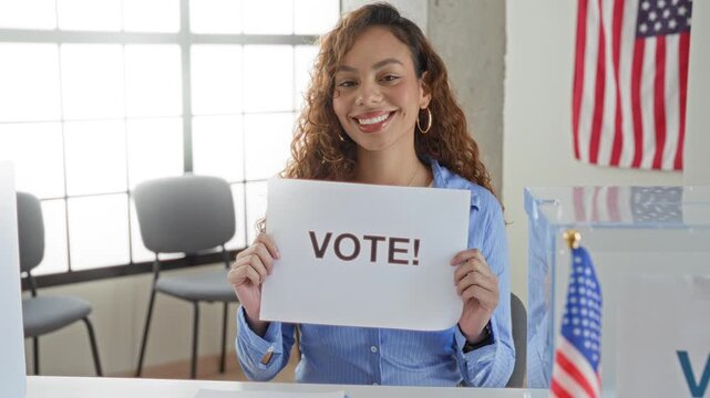Woman holding sign reading vote! with both hands at a polling station beside a clear ballot box and us flag; civic pride.