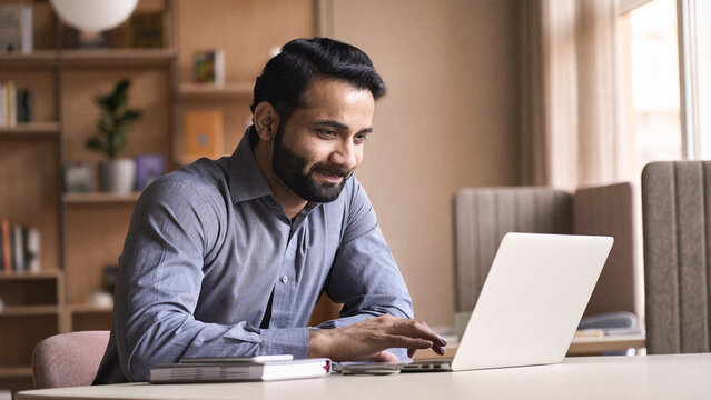 Smiling indian business man working on laptop at home office. Young indian student or remote teacher using computer remote studying, virtual training, watching online education webinar at home office.