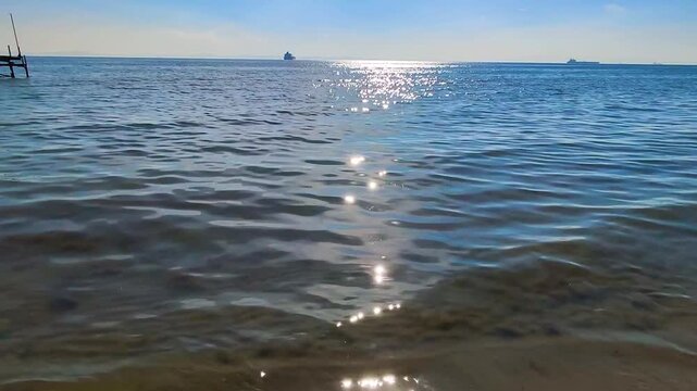 Breathtaking View of Bright Sun Glitters Reflecting on Calm Blue Sea Water, Featuring Distant Cargo Ships on the Horizon and Gentle Waves Lapping the Sandy Shore, Serene Maritime Background