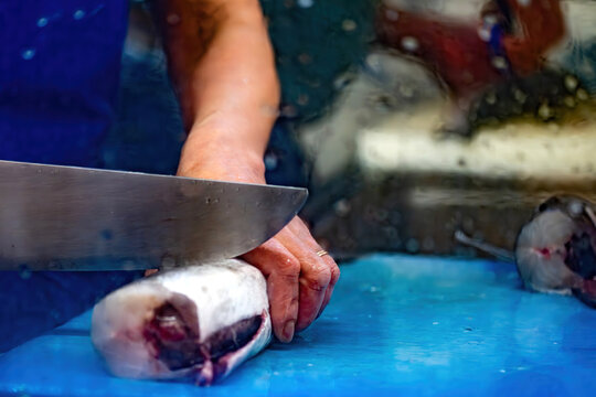 View of a person's hands using a large sharp knife to slice fresh fish on a blue cutting board in a market with a fish head visible in the background.