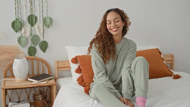 Young latin woman in loungewear sitting on bed with hand on knee smiling, curly hair and pink socks, orange pillows visible in building; cozy comfort serene.