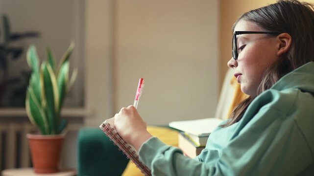 Smart Little Girl in Big Glasses Writing in Notebook while Sitting on Couch with Stack of Books &mdash; Cinematic 4K 10-bit