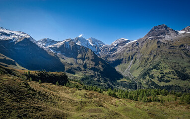 Fototapeta premium Peaceful picturesque mesmerizing atmospheric landscape with high Snow-capped mountains, alpine serpentine pass road, and meadow grasslands pasture with blue sky panorama