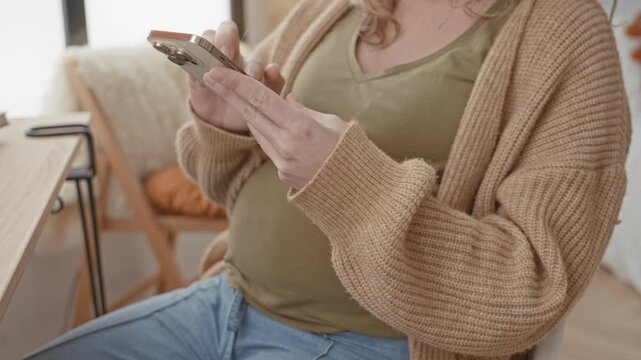 Pregnant woman holding smartphone with hands, texting while seated near a wooden table in a building; calm anticipation.