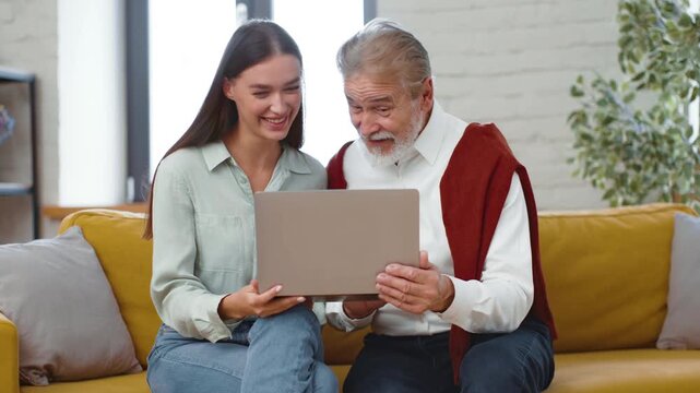 A young woman and an older man sit on a yellow couch, looking at a laptop screen. They smile and interact with the device in a bright room, enjoying a shared moment.