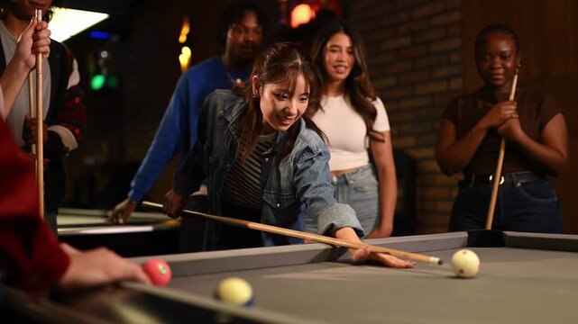Diverse group of friends celebrating with cheers as a joyful woman sinks a ball while playing pool together in a dimly lit bar