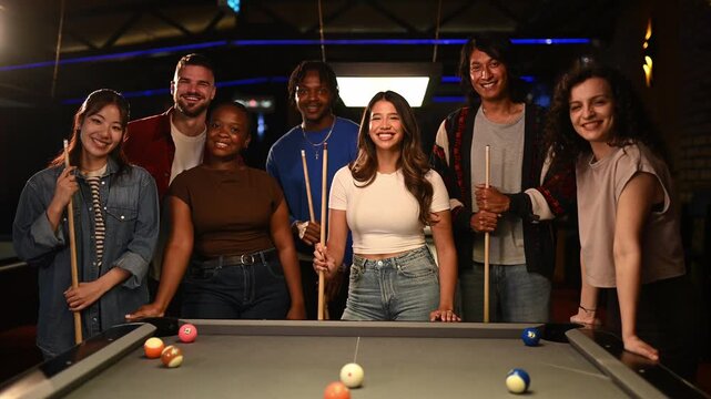 Diverse group of friends smiling and posing together around a pool table holding cues in a modern billiards hall at night