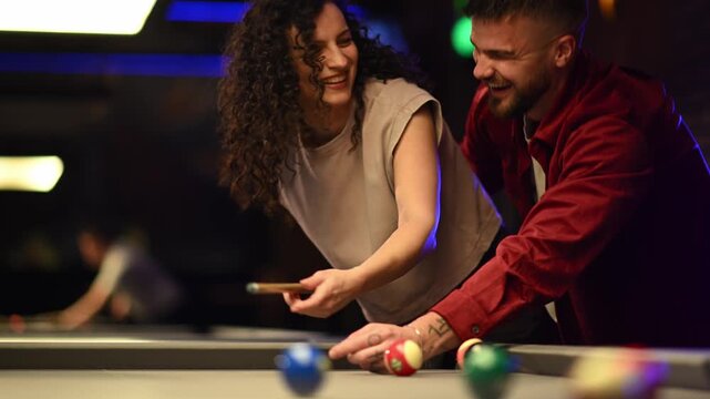 Romantic couple playing billiards in a bar, man teaching his girlfriend how to hold the cue stick and aim on a date night