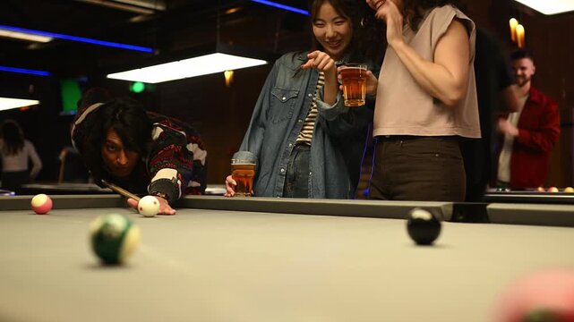 Focused asian man hitting a cue ball playing pool with excited friends watching and cheering in a modern bar