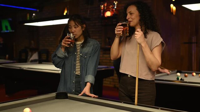 Two women drinking beer and talking while playing pool together in a bar with friends at night