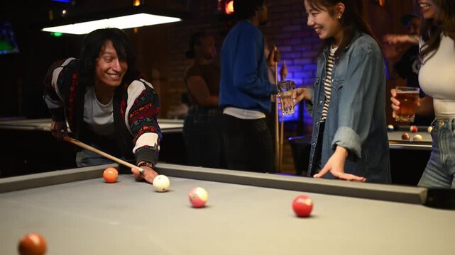 Young multicultural friends laughing and playing billiards together in a modern club at night while enjoying drinks