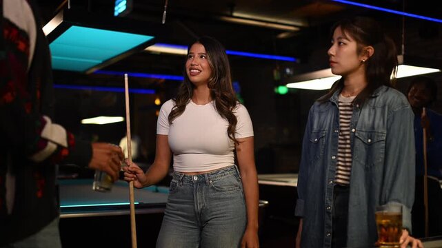 Young latina woman holding a cue stick and chatting with friends while playing billiards in a lively pool hall at night