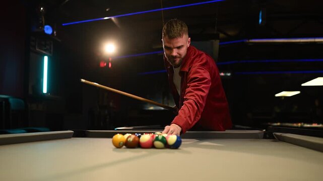 Confident man making a breaking shot while playing pool in a dark, modern billiard club with neon lights