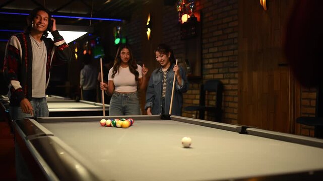 Cheerful young friends having fun and laughing while playing billiards in a modern pool hall at night