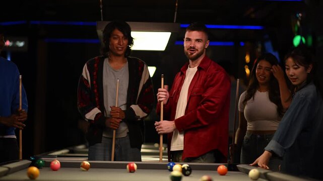 Diverse group of young friends having fun and playing billiards together in a dark, modern pool hall on a friday night