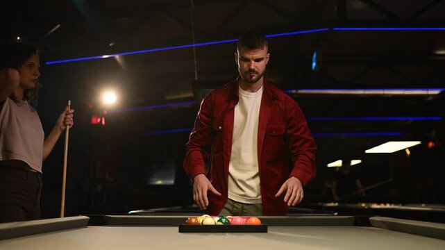 Couple preparing a pool game, man racking the balls on the table in a dark bar with neon lights