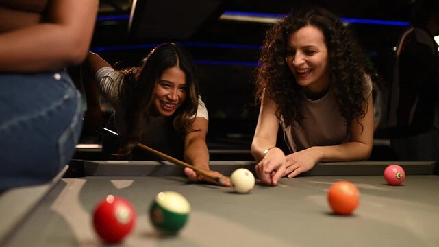Joyful young woman sinking a ball while playing pool with her friends in a dark billiard hall