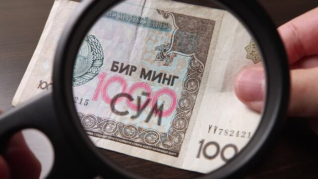 Examination of 1000 Uzbekistan Sum (UZS) cash money with magnfication glass, loupe. Concept of authenticity check, fight against counterfeiting of banknotes, anti fraud measures in Uzbekistan.