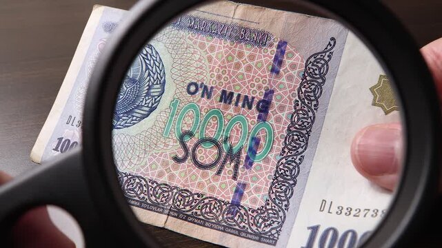 Examination of 10000 Uzbekistan Sum (UZS) cash money with magnfication glass, loupe. Concept of authenticity check, fight against counterfeiting of banknotes, anti fraud measures in Uzbekistan.