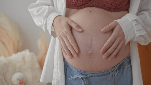 Woman pregnant hands form heart on exposed belly wearing lace bra and loose denim jeans in bedroom with plush pillow and pampas grass; maternal tenderness bonding.