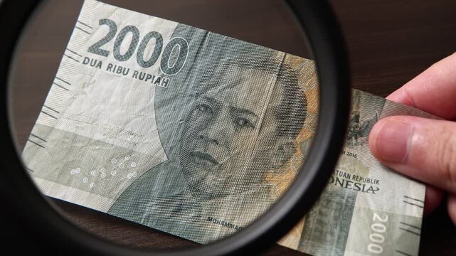 Examination of Indonesian Rupiah cash money with magnfication glass, loupe. Concept of authenticity check, fight against counterfeiting of banknotes, anti fraud measures in Indonesia. Close up view.