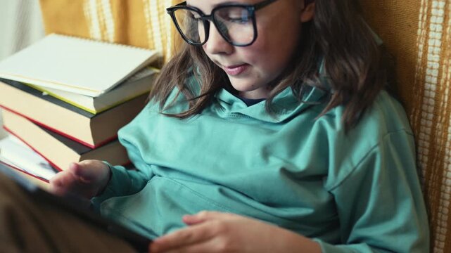 Little Girl in Glasses Using Digital Tablet on Couch with Stack of Books, Child Studying with Pad at Home, 4K 10-bit