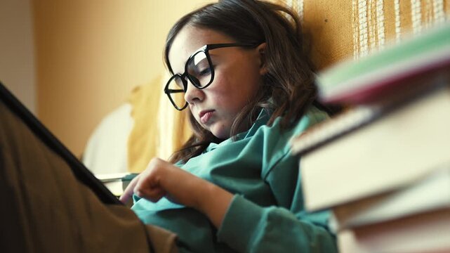 Little Girl in Glasses Using Digital Tablet on Couch with Stack of Books, Child Studying with Pad at Home, 4K 10-bit
