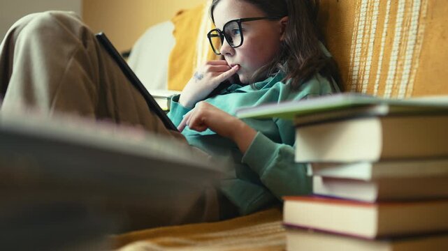 Little Girl in Glasses Using Digital Tablet on Couch with Stack of Books, Child Studying with Pad at Home, 4K 10-bit
