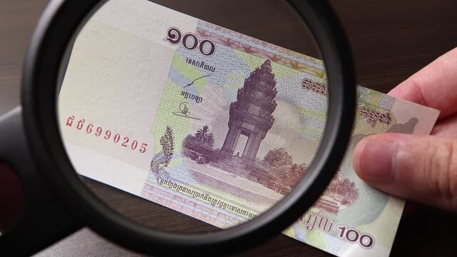 Examination of 100 Cambodian Riel (KHR) cash money with magnfication glass, loupe. Concept of authenticity check, fight against counterfeiting of banknotes, anti fraud measures in Cambodia.