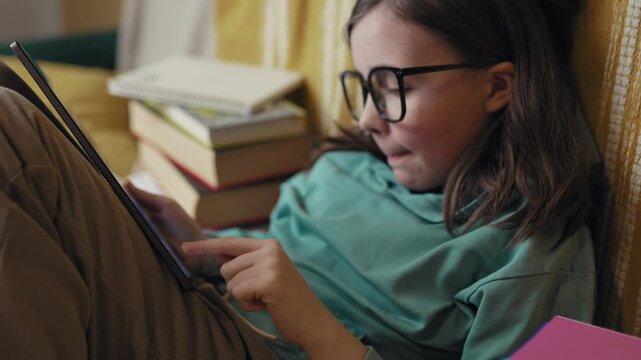Little Girl in Glasses Using Digital Tablet on Couch with Stack of Books, Child Studying with Pad at Home, 4K 10-bit
