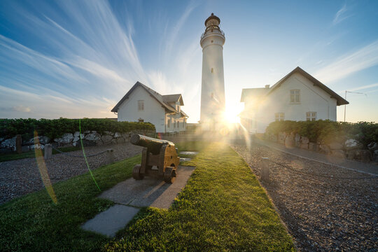 View of Hirtshals fyr lighthouse with a historic cannon in the foreground and white buildings under a sun flare and wispy blue sky Hirtshals, Denmark.